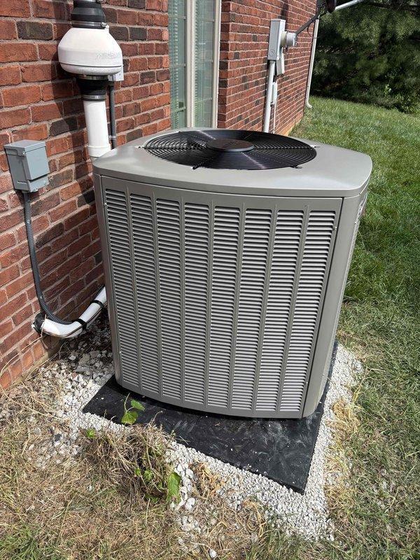 # HVAC Install - Complete System

Successfully completed a full residential HVAC system installation including both indoor and outdoor components. The interior installation features a new furnace unit positioned in the utility area with properly configured white insulated ductwork extending from the unit, secured with metal clamps and routed efficiently across the ceiling space. Installed gray metal plenum with blue gauge monitoring device and yellow flexible coupling connector. All gas and electrical connections completed with black conduit running vertically for proper service access.

The exterior installation includes a new air conditioning condenser unit positioned on a composite pad in the side yard, surrounded by white gravel for drainage and equipment protection. Mounted unit features vertical louvered panels and top-mounted fan assembly. Completed refrigerant line installation with white PVC piping running vertically along the brick exterior wall for proper condensate drainage