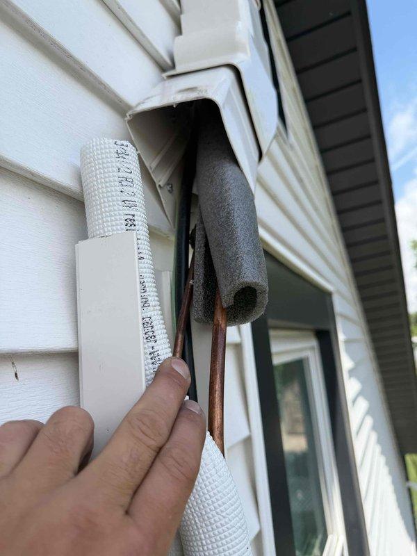 Responded to water leak concern on exterior of residence where condensate drain line was reported to be overflowing and dripping onto window sill. Upon inspection, located condensate drainage piping on side of building beneath eave. Examined refrigerant line set and condensate drain routing from HVAC system. Found deteriorated insulation on copper refrigerant lines with black foam insulation split and separated from piping, exposing underlying insulation material. Assessed condensate drain line installation and verified proper pitch and drainage flow. Inspected mounting brackets and pipe penetrations through building exterior. Confirmed condensate removal system was functioning correctly with no active overflow condition present at time of service. Documented existing condition of exterior piping insulation showing weather deterioration and recommended monitoring for future maintenance needs.