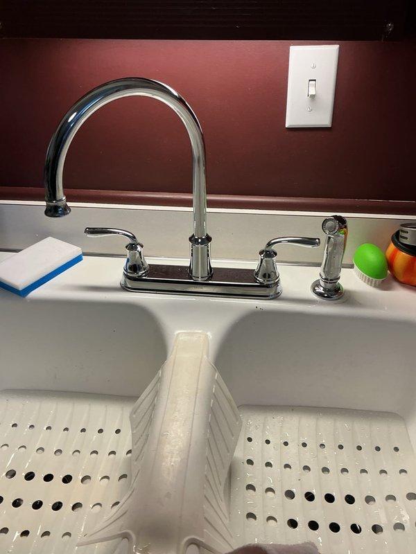 Installed customer-supplied double-handle chrome kitchen faucet with separate spray nozzle attachment on white double-basin sink. Additionally installed 1/2" ball valve on water supply line, connecting blue PVC pipe to flexible braided supply hoses to complete the plumbing installation.