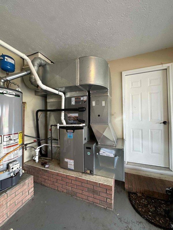 # HVAC Complete System Installation

Successfully completed installation of a 3.5-ton, 15.5 SEER air conditioning system paired with a 97% AFUE gas furnace at residential property. Installed Lennox outdoor condensing unit on concrete pad with proper leveling and clearances. Unit positioned on exterior brick wall with decorative rock landscaping base. Mounted weatherproof electrical disconnect box on exterior wall with appropriate conduit and wiring per code requirements. Routed refrigerant line set through brick wall penetration with proper sealing.

Interior installation completed in utility room with equipment mounted on elevated brick platform for drainage and accessibility. Installed complete air handler system with vertical configuration to maximize space efficiency. Integrated new furnace unit with existing ductwork connections. Mounted humidification system components including metallic cylindrical humidifier assembly near ceiling level. All equipment secured with proper mountin