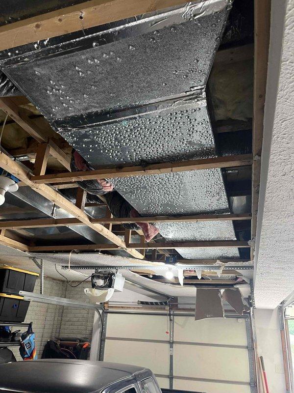 Responded to service call for condensation dripping from main AC unit onto garage floor. Upon inspection, discovered extensive condensation accumulation on exposed ductwork and metallic HVAC components in garage ceiling area. Heavy moisture beading observed across multiple duct panels, indicating significant temperature differential between conditioned air inside ducts and warm, humid garage ambient air. 

Identified cause as lack of proper duct insulation on supply ductwork running through unconditioned garage space. Cold air flowing through uninsulated metal ducts created ideal conditions for atmospheric moisture to condense on exterior duct surfaces. System confirmed operational and cooling effectively; condensation issue isolated to ductwork installation rather than equipment malfunction.

Documented conditions with photographs showing condensation pattern across rectangular metal duct sections and surrounding wooden ceiling framing. Advised that proper remediation requires insulat
