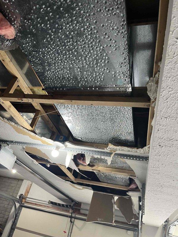 Responded to service call for condensation dripping from main AC unit onto garage floor. Upon inspection, discovered extensive condensation accumulation on exposed ductwork and metallic HVAC components in garage ceiling area. Heavy moisture beading observed across multiple duct panels, indicating significant temperature differential between conditioned air inside ducts and warm, humid garage ambient air. 

Identified cause as lack of proper duct insulation on supply ductwork running through unconditioned garage space. Cold air flowing through uninsulated metal ducts created ideal conditions for atmospheric moisture to condense on exterior duct surfaces. System confirmed operational and cooling effectively; condensation issue isolated to ductwork installation rather than equipment malfunction.

Documented conditions with photographs showing condensation pattern across rectangular metal duct sections and surrounding wooden ceiling framing. Advised that proper remediation requires insulat