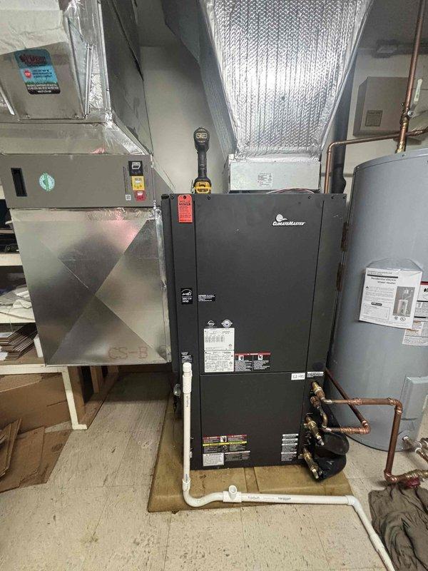 Completed installation of geothermal HVAC system including black air handler unit with integrated components and gray water heater. Connected copper piping with brass fittings and valves between units, installed white PVC condensate drain line, and completed silver reflective ductwork with proper insulation and plenum transitions in mechanical room with tile flooring.
