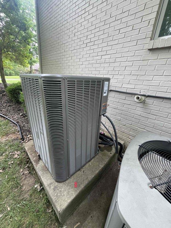 Completed installation of a 5-ton, 14.7 SEER air conditioning system with matching evaporator coil at residential property. The outdoor condenser unit was positioned on a concrete pad against the exterior white brick wall with proper electrical conduit routing and refrigerant line penetration through the wall. The indoor evaporator coil was integrated with the existing Lennox furnace system in the basement utility area, with appropriate condensate drain lines and PVC venting installed to ensure proper system operation and code compliance.