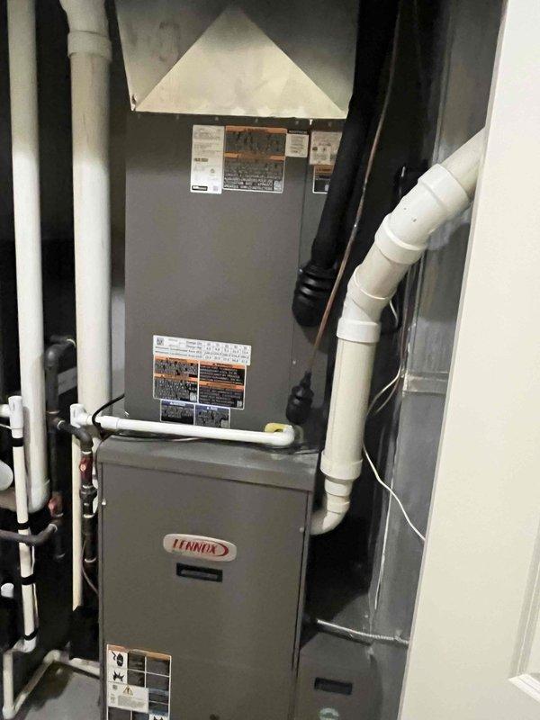 Completed installation of a 5-ton, 14.7 SEER air conditioning system with matching evaporator coil at residential property. The outdoor condenser unit was positioned on a concrete pad against the exterior white brick wall with proper electrical conduit routing and refrigerant line penetration through the wall. The indoor evaporator coil was integrated with the existing Lennox furnace system in the basement utility area, with appropriate condensate drain lines and PVC venting installed to ensure proper system operation and code compliance.