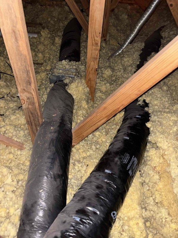 Responded to water leak complaint in utility closet housing 4-year-old Goodman HVAC system and AO Smith water heater. Conducted thorough inspection of attic space and identified improperly installed flexible ductwork routing through ceiling joists with inadequate sealing at roof penetration points. Black flexible ducts showed signs of moisture intrusion and improper slope allowing condensation accumulation. Traced water entry point to compromised vent flashing connection at roof line where HVAC exhaust penetrates roofing structure. Confirmed roofer's assessment that ductwork installation did not follow proper building codes for attic ventilation systems. Water damage evident on utility closet flooring required placement of air movers to prevent further deterioration. Documented attic conditions showing loose-fill fiberglass insulation, exposed ceiling joists, and three runs of flexible ductwork with visible wear and dust accumulation. Provided recommendations for proper duct reconfigur
