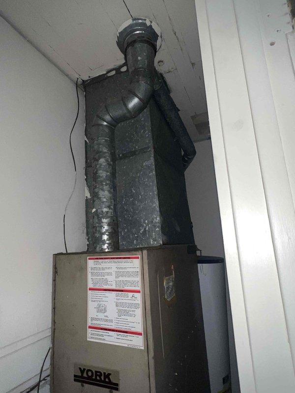 Performed comprehensive assessment of existing residential HVAC system for replacement estimate. Evaluated 40-year-old air conditioning unit located exterior to residence, documenting original equipment from 1980s-1990s era that has reached end of serviceable life. Inspected York furnace installation in interior utility space, noting condensation accumulation on vertical plenum component and flexible ductwork connections. Photographed existing equipment configuration including indoor air handler placement in confined utility area with wooden enclosure and outdoor condensing unit location accessible via rear alley approach. Documented current system specifications, installation conditions, and spatial constraints to provide accurate replacement equipment recommendations and installation requirements for complete air conditioning system upgrade.