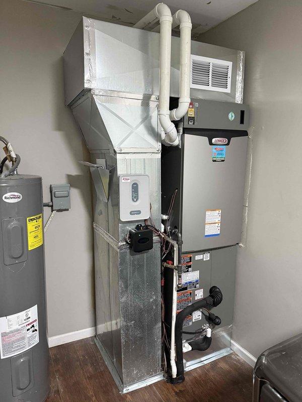 # HVAC Complete System Installation

Successfully completed installation of a 4-ton, 17 SEER zoned HVAC system with 97% AFUE heating capability. The project included both indoor and outdoor equipment installation with proper refrigerant line routing and electrical connections.

## Indoor Installation

Installed air handler unit with metallic silver finish in utility area, positioned vertically against interior wall. Mounted large white pyramidal plenum chamber above air handler to facilitate proper airflow distribution throughout the ductwork system. Ran dual white PVC condensate drain lines vertically alongside equipment to ensure proper drainage and prevent water damage. Installed wall-mounted white rectangular air return vent for adequate system circulation.

Connected dark-colored heating unit with exposed copper refrigerant lines, properly insulated with black coating. Completed all electrical connections and verified proper component integration. Ensured all manufacturer labels a