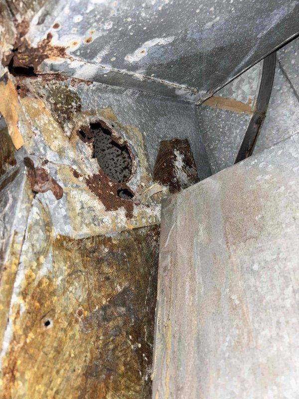 Responded to no-cooling call on 5-6 year old Carrier system with indoor temperature reading 79°F. Upon arrival, confirmed outdoor condenser fan motor was inoperative while compressor hummed, indicating failed condenser fan motor. Inspection of outdoor unit revealed severely deteriorated ductwork with extensive rust corrosion and structural failure, including large hole penetration in metal housing compromising system integrity. Documented equipment condition showing advanced oxidation and moisture damage throughout condensing unit cabinet and associated components.