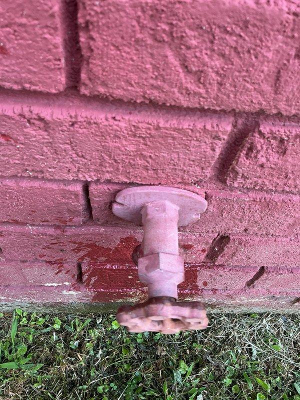 Responded to service call for non-functional outdoor hose spigot attached to exterior wall and assisted with locating main water shut-off valve. Upon inspection, identified outdoor spigot fixture mounted on pink brick exterior wall at ground level. Successfully located main water shut-off valve in crawl space access point beneath structure, identifying blue-handled shut-off valve on water supply line along with associated PVC piping and plumbing infrastructure. Provided estimate for installation of interior shut-off valve as alternative to existing exterior yard valve location that requires access outside property fence line.