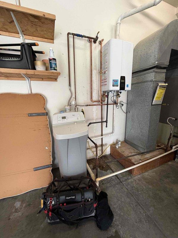 Completed professional installation of tankless water heater system in residential utility area. Installation included proper mounting of unit to wall, connection of copper supply lines, integration with existing water softener system, installation of pressure relief valve with appropriate safety components, electrical wiring with dedicated circuit and grounding, and flexible gas line connection using corrugated metallic tubing. All plumbing connections were secured with code-compliant fittings, electrical components were properly terminated and protected, and unit was tested for proper operation with digital display confirming system functionality.
