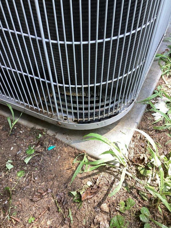# HVAC Maintenance Summary

Completed Platinum Level preventive maintenance on single outdoor condensing unit. Performed comprehensive system inspection including visual assessment of condenser coil, protective grille, and mounting pad. Documented unit condition with photographic evidence showing exterior housing with vertical slat grille design and cylindrical configuration. 

Noted vegetation encroachment around concrete pad perimeter with weeds and grass growing near unit base. Observed normal weathering on exterior components including dirt accumulation and discoloration on lower portion of protective grille. Concrete mounting pad showed age-appropriate weathering with moisture staining. Internal components visible through grille appeared intact with no obvious damage to fins or coil structure.

System operational at time of inspection with no immediate mechanical concerns identified. Unit positioned on stable concrete foundation with adequate clearance for airflow. Recommended veg