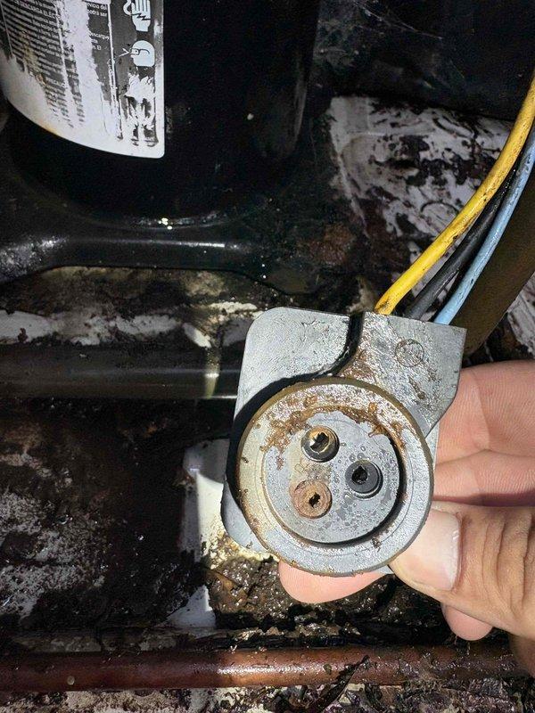 Performed maintenance inspection on 20-year-old electric air conditioning system. Upon examination, discovered severely corroded reversing valve and drain pan with extensive rust buildup and mineral deposits. Component showed significant deterioration with brownish-orange discoloration around valve ports and granular flaking on metal surfaces. Documented conditions with photographs showing compromised equipment integrity, white mineral efflorescence, and moisture damage throughout the attic installation. Advised customer of findings and recommended replacement of affected components due to advanced corrosion that could lead to system failure or refrigerant leaks.