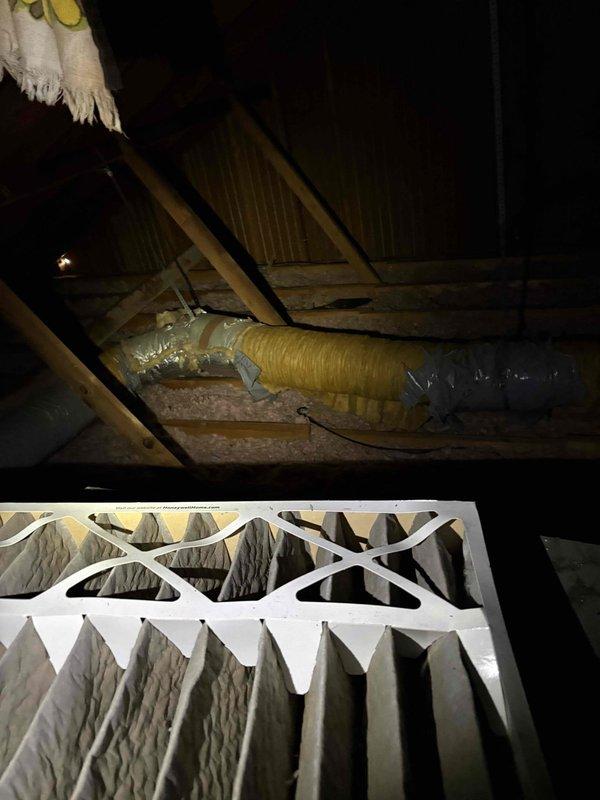 Responded to service call for air conditioning system blowing warm air at vacant property. Upon arrival, accessed property using lockbox and confirmed system had been turned off by occupants. Performed diagnostic inspection of HVAC system including attic-mounted ductwork and air handler components. Inspected flexible ductwork runs throughout attic space, verifying proper installation, support, and insulation integrity. Examined air filtration system and confirmed clean filter installation in return air housing. Documented all accessible system components including ductwork routing, connections, insulation wrapping, and structural mounting points. Photographed attic installation showing yellow flexible supply ducts, pink fiberglass insulation near roof decking, and multiple gray insulated duct runs secured to wooden framing members. System requires further evaluation to determine root cause of warm air discharge issue.