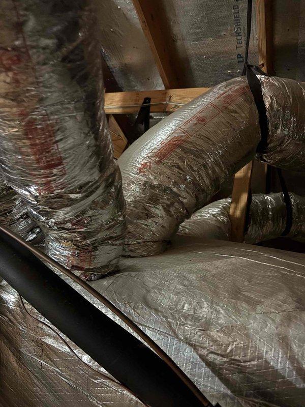 Responded to after hours service call for air conditioning system experiencing repeated freeze-up conditions. Upon arrival, unit was powered off with only the air handler fan operating. Performed diagnostic inspection of HVAC system installed in attic space. Examined air handler unit, flexible ductwork installation, and associated components. Documented system configuration including metallic flexible ducts properly secured with metal straps to wooden framing, insulated air handler panels, and copper refrigerant lines. Assessed overall condition of ductwork insulation and verified proper installation methods. System appeared to be approximately ten years old based on visual inspection. Captured photographic documentation of attic-mounted equipment, duct routing, and installation details for service records.