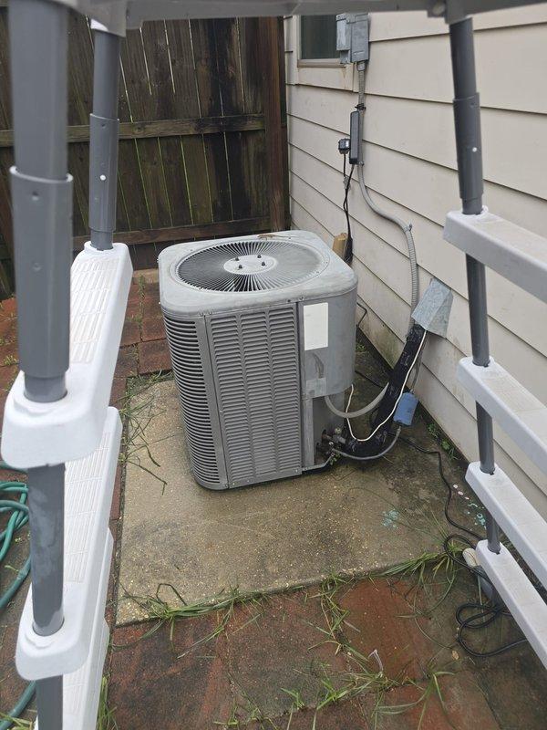 Completed condenser unit changeout at residential property. Removed existing outdoor air conditioning condenser and installed new replacement unit on existing concrete pad in exterior utility area between building walls. Verified proper leveling of unit on pad to ensure optimal performance and drainage. Established electrical connections from weatherproof junction box mounted on building exterior wall to new condenser via conduit. Confirmed proper placement and stability of unit on weathered concrete pad surface. Documented installation with photographs of installed unit showing positioning on concrete pad with surrounding brick paver area, electrical connections, and placement adjacent to building siding. Captured required documentation photographs of equipment data tag displaying model number and serial number information for warranty registration and service records.