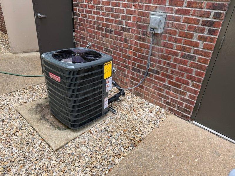 Completed installation of 2-ton split system HVAC unit including outdoor condenser and indoor air handler. Condenser unit positioned on concrete pad against exterior brick wall with proper clearance and secured electrical disconnect. Air handler installed in basement utility area with appropriate ductwork connections and condensate drainage. All electrical conduit properly routed and secured to building exterior. System installation includes manufacturer labeling, safety warnings, and operational specifications clearly visible on equipment. Unit tested and verified operational upon completion.