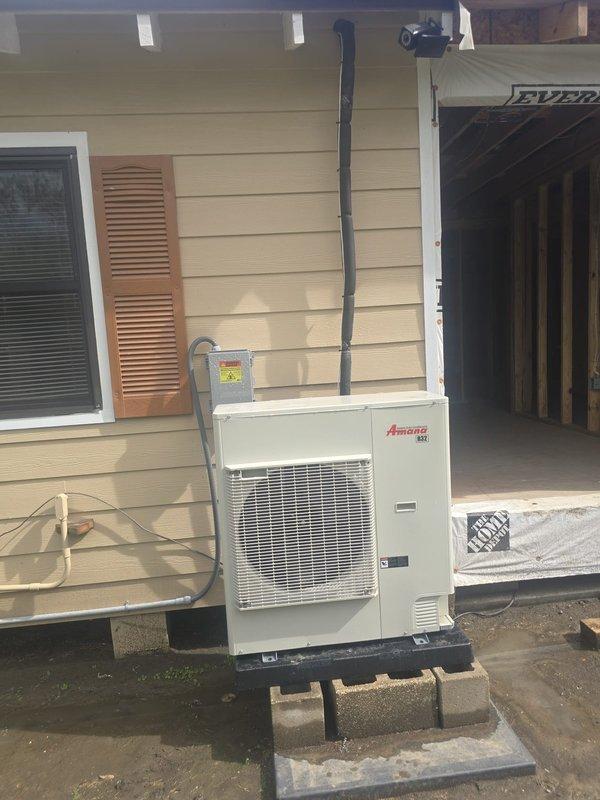 Conducted site assessment of existing Amana outdoor HVAC condenser unit to evaluate feasibility of relocation. Documented current installation configuration, including unit placement on concrete block platform, refrigerant line routing along exterior wall, and electrical conduit connections extending to roofline. Photographed existing setup showing proximity to window with shutters and adjacent garage area to determine clearance requirements and potential relocation options for improved placement or accessibility.