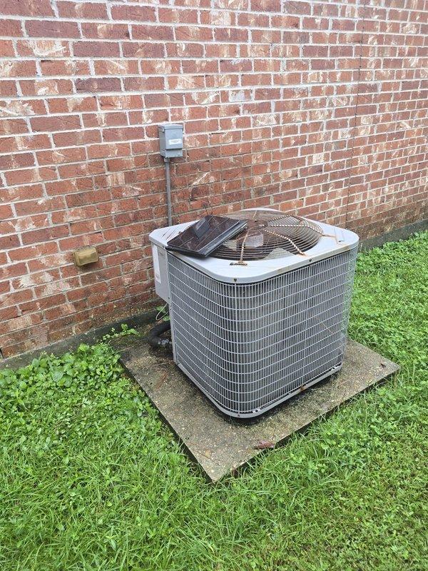 Responded to service call for non-operational air conditioning condenser unit. Upon arrival, inspected existing outdoor HVAC system positioned on concrete pad against exterior brick wall. Unit features gray metal construction with protective grille covering and electrical disconnect box mounted above with conduit connections. Assessed current condenser condition and surrounding installation components including concrete pad, electrical connections, and refrigerant line routing. Provided estimate for complete condenser replacement as requested, excluding full system replacement options per customer preference.