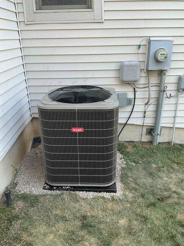 Completed installation of Bryant brand air conditioning condenser unit on exterior of residential property. Unit was positioned on black protective pad over white gravel base against home's vinyl siding, with proper clearance from structure for adequate airflow and service access. Electrical connections were established through existing meter assembly and junction boxes mounted on exterior wall, with conduit runs secured vertically along siding per code requirements. System was placed on level gravel foundation to ensure proper drainage and equipment stability, with all electrical disconnects and grounding components properly installed for safe operation and future maintenance access.