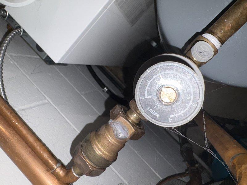 Responded to service call for boiler leak originating from bypass piping on rear of unit. Upon inspection, identified active water leak from corroded copper bypass pipe with visible oxidation and deterioration at connection points. Observed pressure gauge and associated fittings showing signs of age and wear. Water had accumulated on floor despite main water supply being shut off to residence, indicating residual pressure in system. Documented condition of affected copper piping, brass fittings, and pressure monitoring equipment with photographic evidence showing extent of corrosion and leak source location.