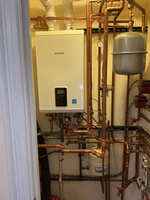 Completed installation of wall-mounted Navien combi boiler system in residential mechanical room. Installation included comprehensive copper piping network with multiple shut-off valves, gray expansion tank, white PVC venting, and integrated water filtration component. System was professionally assembled with proper valve placement, Energy Star certified unit, and digital control panel for efficient hydronic heating operation.