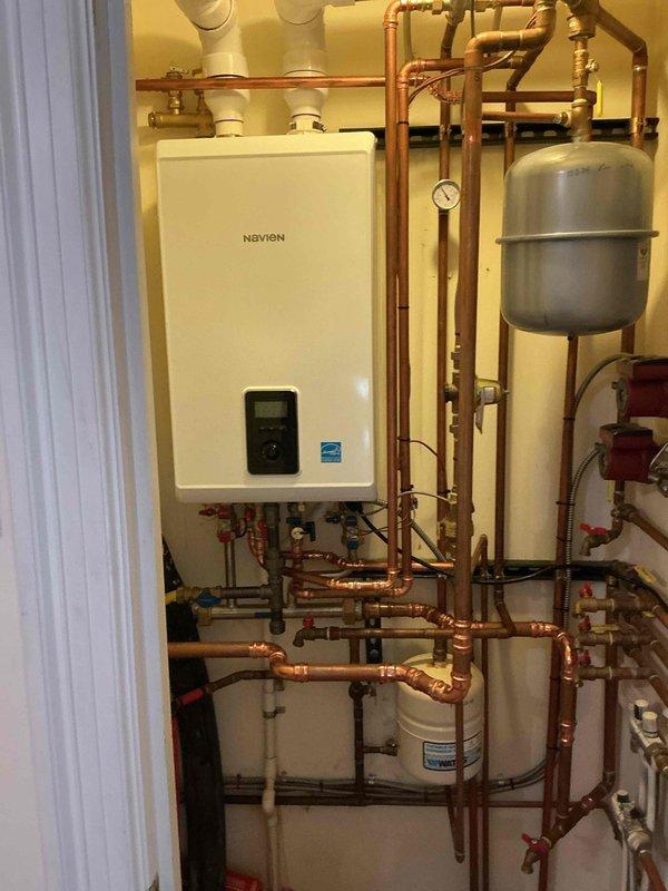 Completed installation of wall-mounted Navien combi boiler system in residential mechanical room. Installation included comprehensive copper piping network with multiple shut-off valves, gray expansion tank, white PVC venting, and integrated water filtration component. System was professionally assembled with proper valve placement, Energy Star certified unit, and digital control panel for efficient hydronic heating operation.