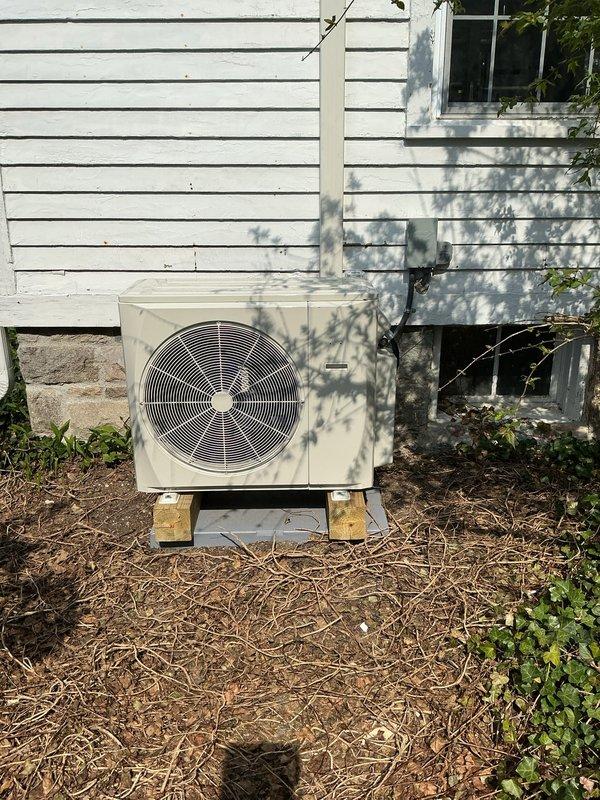 Completed installation of multizone mini split system including outdoor condenser unit mounted on wooden blocks with concrete pad and indoor air handler with ductwork in attic space. Refrigerant lines and electrical conduit were run vertically along exterior siding, and all mechanical equipment was properly secured with control panel installed and operational.