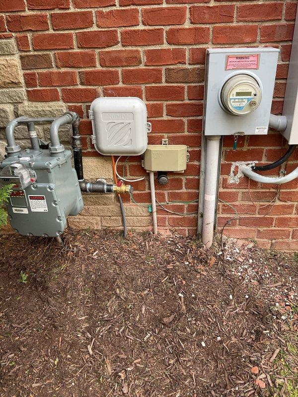 Completed installation of Honeywell residential generator system including exterior utility connections, gas line piping with proper mounting brackets and flexible conduit routing along brick exterior wall, electrical meter integration, and junction box installation. All components were professionally secured to building exterior with appropriate weatherproofing and code-compliant conduit work connecting gas service to generator location.