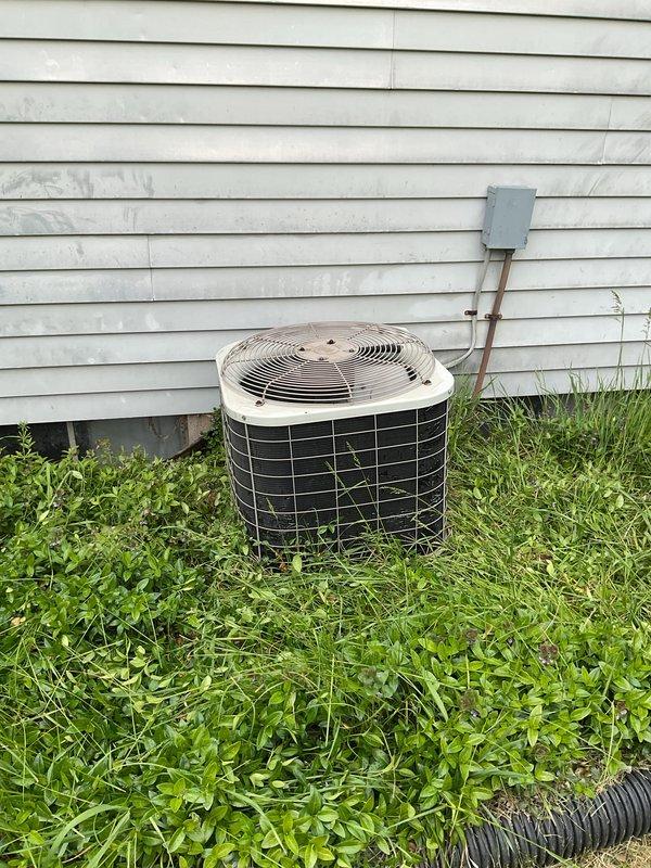 Performed comprehensive AC tune-up on outdoor condenser unit. Inspected cylindrical condenser with beige fan guard and black metal grille cage, verified proper operation of fan motor and electrical connections at disconnect box, and examined copper refrigerant lines running to building. Cleared overgrown grass and vegetation from around unit base to restore proper airflow clearance and prevent debris accumulation that could impact system efficiency.