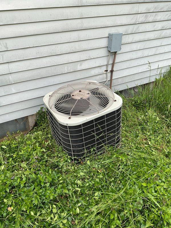 Performed comprehensive AC tune-up on outdoor condenser unit. Inspected cylindrical condenser with beige fan guard and black metal grille cage, verified proper operation of fan motor and electrical connections at disconnect box, and examined copper refrigerant lines running to building. Cleared overgrown grass and vegetation from around unit base to restore proper airflow clearance and prevent debris accumulation that could impact system efficiency.