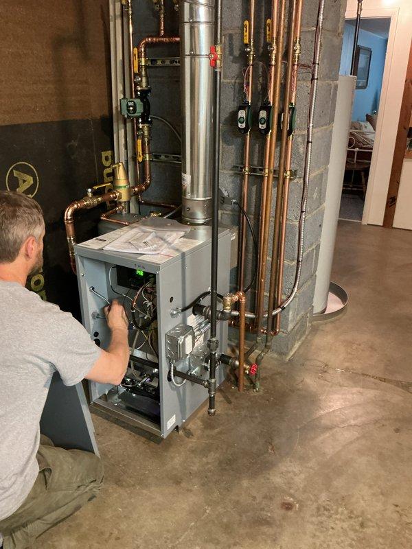 Completed installation of new residential boiler and hot water heater in basement utility area. Technician accessed and configured internal burner assembly and mechanical components within the boiler unit, ensuring proper operation and system integration. Installed and connected copper piping with brass fittings and shut-off valves with red handles along concrete block wall for supply and return lines. Properly vented system with silver flue pipe extending vertically from boiler unit. Installed tan-colored residential water heater with associated copper piping connections and blue pressure tank for water treatment system. All mechanical systems tested and verified operational upon completion.