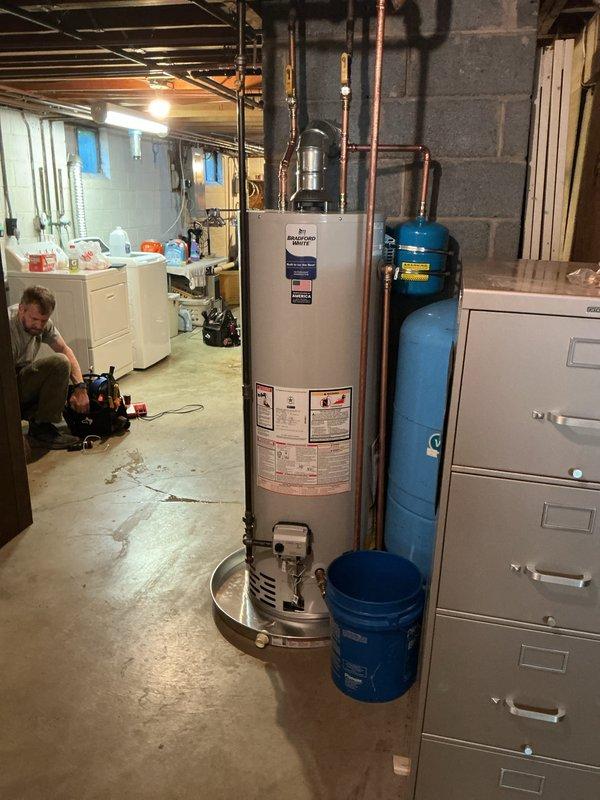 Completed installation of new residential boiler and hot water heater in basement utility area. Technician accessed and configured internal burner assembly and mechanical components within the boiler unit, ensuring proper operation and system integration. Installed and connected copper piping with brass fittings and shut-off valves with red handles along concrete block wall for supply and return lines. Properly vented system with silver flue pipe extending vertically from boiler unit. Installed tan-colored residential water heater with associated copper piping connections and blue pressure tank for water treatment system. All mechanical systems tested and verified operational upon completion.
