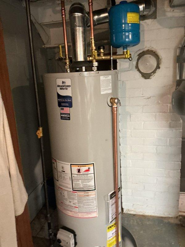 Completed installation of a Bradford White automatic storage water heater in residential basement utility area. Installed new unit with proper plumbing connections including copper piping, brass fittings, flexible metal connectors, and blue expansion tank mounted horizontally above the water heater. All connections secured to manufacturer specifications with appropriate safety labels and warning stickers visible on unit exterior.