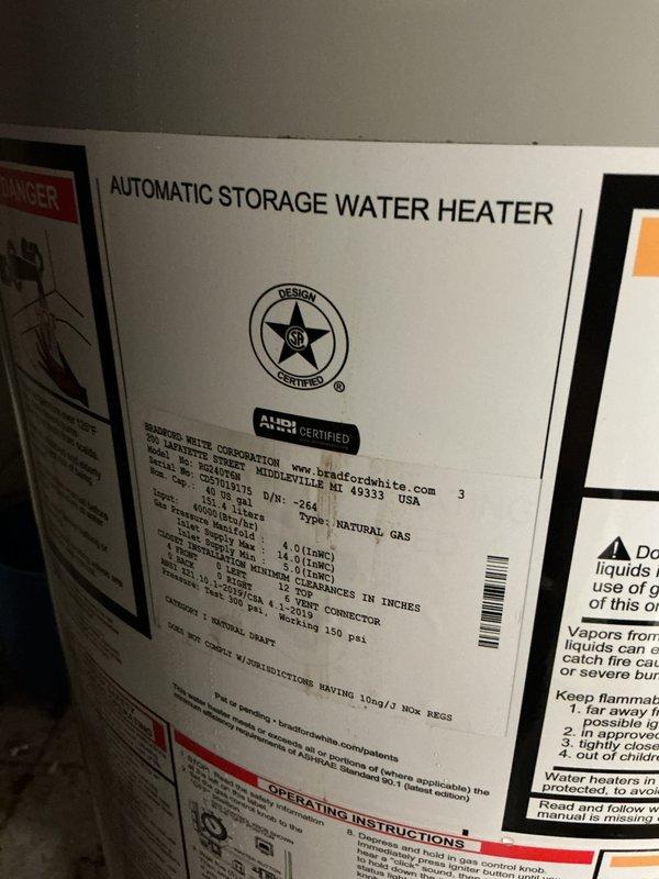 Completed installation of a Bradford White automatic storage water heater in residential basement utility area. Installed new unit with proper plumbing connections including copper piping, brass fittings, flexible metal connectors, and blue expansion tank mounted horizontally above the water heater. All connections secured to manufacturer specifications with appropriate safety labels and warning stickers visible on unit exterior.