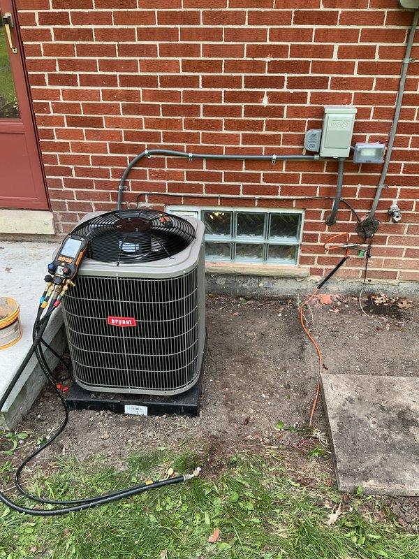Completed installation of Bryant air conditioning system with outdoor condensing unit positioned on concrete pad against brick exterior wall. Unit was properly mounted, refrigerant lines were run vertically along brick wall with secure mounting brackets, electrical disconnect box was installed, and system was charged and tested using manifold gauges to verify proper refrigerant pressures and operational performance.