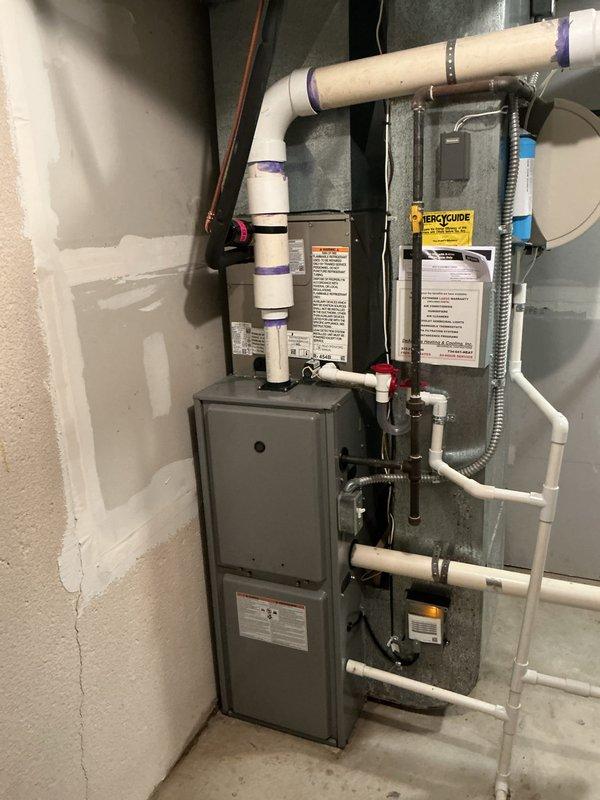 Completed installation of new air conditioning system with evaporator coil in basement utility area. System includes wall-mounted air handler with proper condensate drainage via PVC piping to floor drain, copper refrigerant lines, and integrated ductwork connections to existing furnace. All equipment mounted securely to concrete wall with appropriate clearances, manufacturer labels visible, and system tagged for identification.