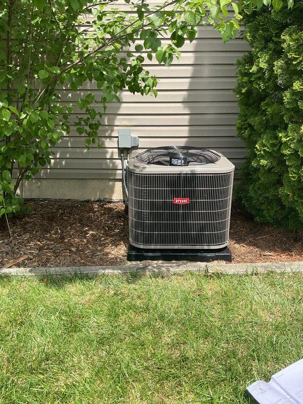 Completed full air conditioning system installation at residential property. Installed new outdoor condensing unit on concrete pad with proper clearances from structure and landscaping. Unit positioned on black platform base against house exterior with adequate airflow access. Connected refrigerant lines and electrical service to new condenser. Interior installation included integration with existing furnace system, routing of refrigerant lines through basement mechanical room, and installation of condensate drainage piping. Verified all ductwork connections at air handler, secured PVC condensate drain lines with proper pitch and trap configuration. Tested system startup, confirmed proper refrigerant charge, verified temperature differential across coil, and ensured all electrical connections meet code requirements. System operating within manufacturer specifications upon completion.