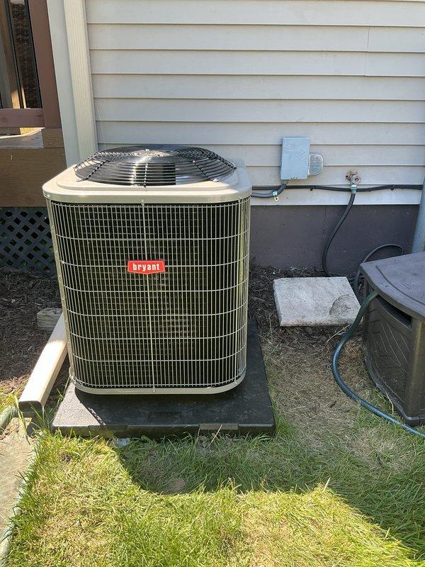 Completed installation of new Bryant air conditioning system with matching evaporator coil at residential property. Unit positioned on concrete pad with proper elevation supports against exterior wall with white vinyl siding. Connected electrical disconnect box with appropriate conduit and wiring to meet code requirements. System installed with protective wire mesh grille covering condenser coils and fan guard on top of unit. All refrigerant lines properly connected and insulated. Electrical connections completed and verified. Unit tested for proper operation and airflow. Work area cleaned and debris removed from site upon completion.