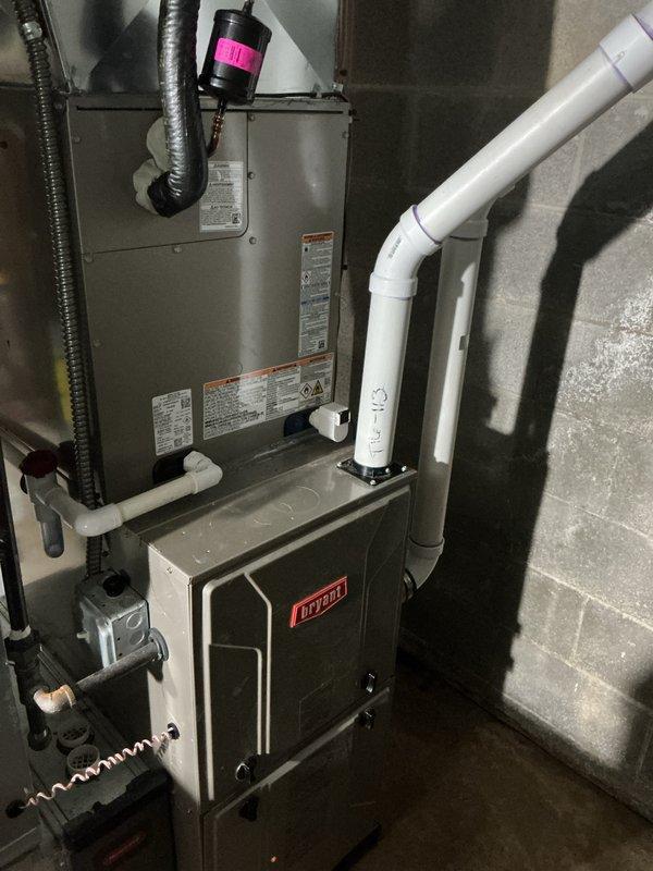 Completed installation of new Bryant HVAC system including air conditioning unit and evaporator coil in residential basement utility room. Mounted air handler unit on concrete block wall above existing furnace, installed refrigerant lines with proper insulation, and connected white PVC condensate drain piping with appropriate trap configuration. Integrated system with existing ductwork infrastructure, verified all electrical connections, and ensured proper clearances for maintenance access. System tested and operational.