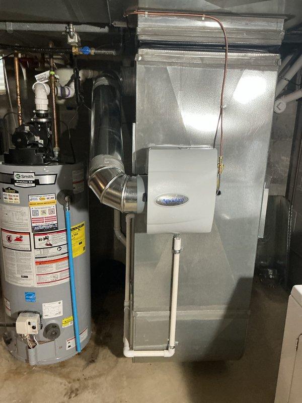 Completed installation of new Bryant HVAC system including air conditioning unit and evaporator coil in residential basement utility room. Mounted air handler unit on concrete block wall above existing furnace, installed refrigerant lines with proper insulation, and connected white PVC condensate drain piping with appropriate trap configuration. Integrated system with existing ductwork infrastructure, verified all electrical connections, and ensured proper clearances for maintenance access. System tested and operational.