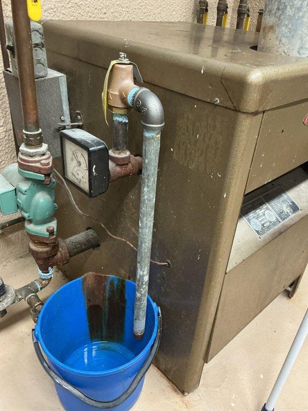 Responded to service call for water leak at boiler system. Upon inspection, identified heavily corroded bolt and fitting assembly on pipe connection near boiler unit with extensive rust damage from prolonged moisture exposure. Leak was actively dripping into collection bucket placed beneath affected area. Assessed deteriorated galvanized piping and compromised connection integrity due to oxidation. Documented existing conditions including rusted components and aging plumbing infrastructure for repair recommendations.