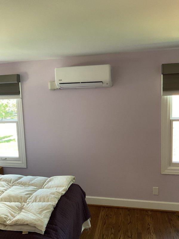Completed installation of ductless mini-split heat pump system in residential bedroom. Mounted indoor air handler unit on interior wall with proper clearances between two windows, installed outdoor condenser unit on exterior brick wall with secure mounting and proper refrigerant line routing through black flexible conduit, and tested system operation to verify proper heating and cooling functionality.