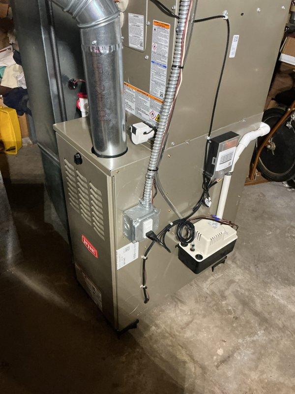 Completed full HVAC system installation including new furnace and air conditioner unit in residential basement utility area. Installed beige vertical furnace unit on concrete pad with proper electrical connections via junction box and control module. Configured metallic supply ductwork from furnace plenum with Y-junction branching system secured to concrete block wall using metal strapping and support hangers. All duct connections fastened with metal bands and clamps per manufacturer specifications. Verified all safety labels properly affixed to equipment and confirmed system ready for startup and commissioning.