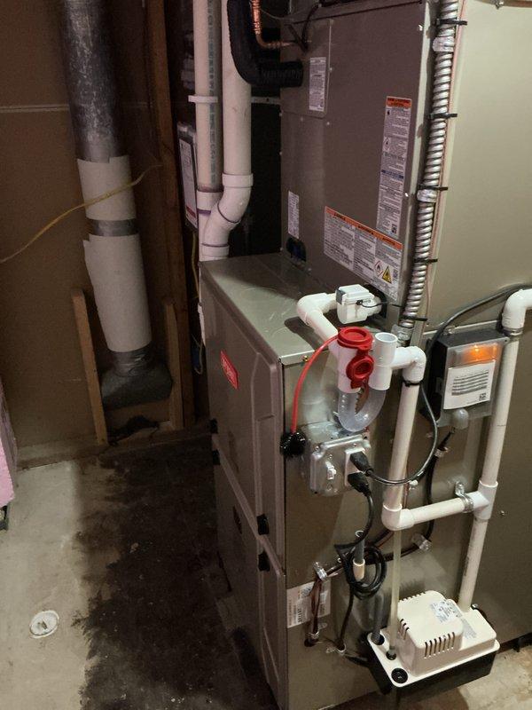Completed full HVAC system installation including new furnace in basement mechanical room and outdoor condensing unit. Installed gray metal furnace with proper venting using white PVC piping, integrated condensate pump with clear reservoir for drainage management, and connected all necessary ductwork including flexible ducting. Mounted outdoor beige condenser unit on black platform pad against brick exterior wall, ran electrical conduit from unit to building, and installed refrigerant lines between indoor and outdoor components. All equipment properly labeled, secured, and tested for operational functionality upon completion.