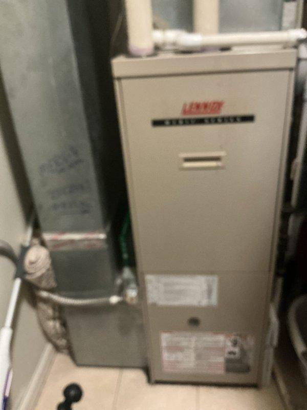 Documented existing HVAC installation featuring a Lennox Model 70E60-6 furnace unit with associated components. Photographed the complete system setup including the beige Lennox furnace positioned beneath a window, gray cylindrical expansion tank with connected piping, gas valve assembly with multiple connection ports, and ductwork configuration. Captured manufacturer specification labels and model identification plates for equipment record purposes.