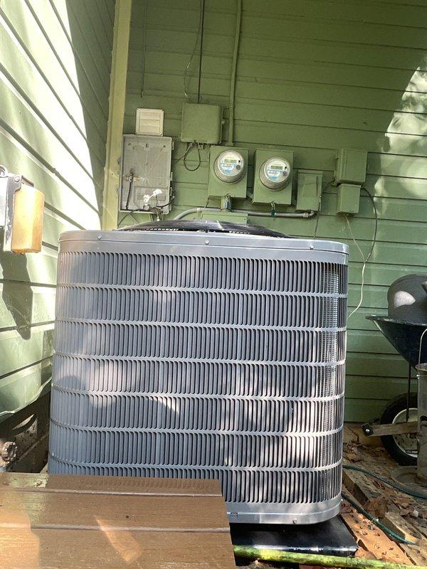 Completed scheduled maintenance tune-up on residential air conditioning system and tankless water heater. Performed comprehensive inspection of outdoor cylindrical condenser unit, verified proper operation of heat exchanger coils and fan assembly, and documented equipment condition with photographs showing unit mounted on wooden platform against exterior wall with associated electrical components including meters and disconnect panel.
