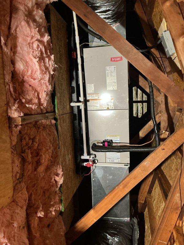 Completed installation of new Bryant HVAC system including air handler and outdoor condensing unit. The air handler was mounted vertically in the attic space between wooden framing members with proper clearance for serviceability. Flexible insulated ductwork was connected to the unit, and all electrical connections were terminated per manufacturer specifications. The outdoor condensing unit was positioned on a concrete pad against the exterior wall with appropriate clearances maintained. Gray electrical conduit and junction boxes were installed along the siding to provide power to the equipment. Refrigerant lines were run from the outdoor unit through the wall to connect with the indoor air handler. All components were properly secured, and the system was tested for proper operation upon completion.