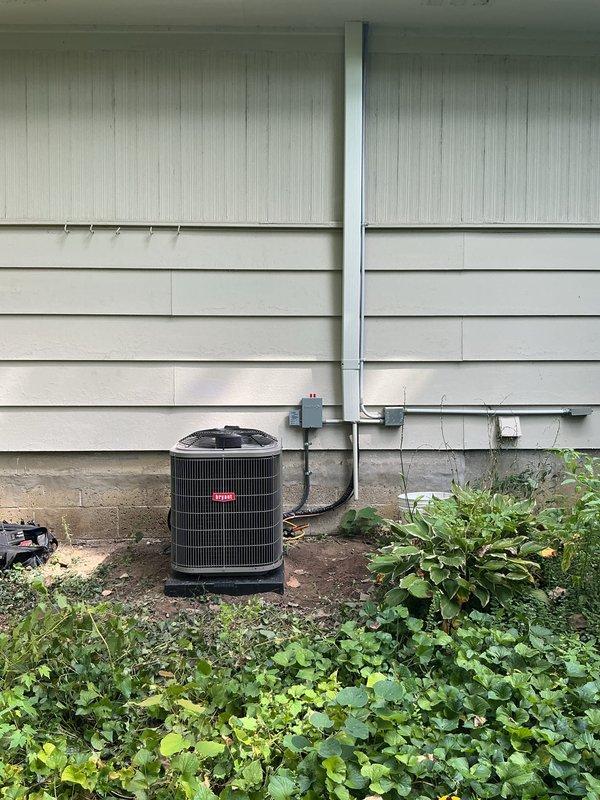 Completed installation of new Bryant HVAC system including air handler and outdoor condensing unit. The air handler was mounted vertically in the attic space between wooden framing members with proper clearance for serviceability. Flexible insulated ductwork was connected to the unit, and all electrical connections were terminated per manufacturer specifications. The outdoor condensing unit was positioned on a concrete pad against the exterior wall with appropriate clearances maintained. Gray electrical conduit and junction boxes were installed along the siding to provide power to the equipment. Refrigerant lines were run from the outdoor unit through the wall to connect with the indoor air handler. All components were properly secured, and the system was tested for proper operation upon completion.