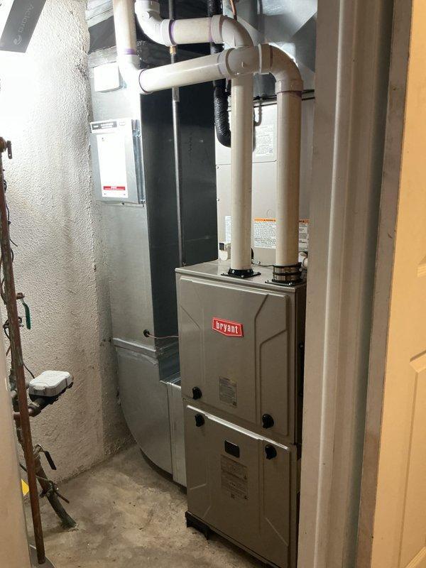 Completed full HVAC system installation including Bryant furnace, outdoor air conditioning condenser unit, and humidifier. Positioned exterior condenser on black pad against foundation with proper clearance, ran refrigerant lines and electrical connections vertically along siding to interior equipment. Interior installation features furnace with dual white PVC intake and exhaust venting routed through ceiling, integrated humidifier system, and proper copper line connections. All equipment professionally mounted with appropriate spacing, ductwork connections secured, and system tested for operational performance.